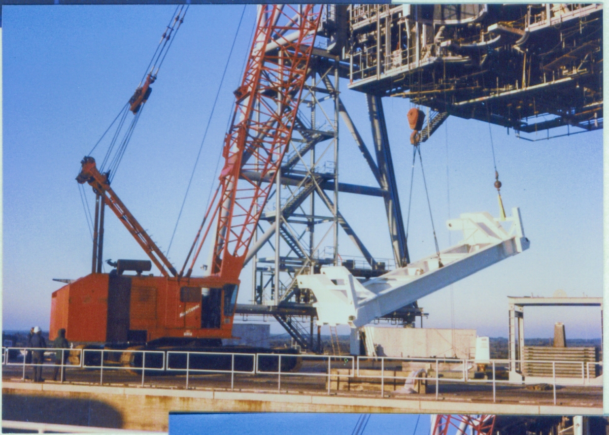 The PGHM Bridge Beam rises into the air, above the pad deck at Launch Complex 39-B, Kennedy Space Center, Florida, on its way to its final location inside the PCR, on the RSS, which is mostly out of frame, extending up and away from the top right corner of the image.